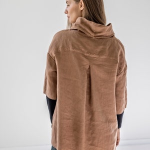 May include: A brown linen shirt with a high collar and three-quarter sleeves. The shirt has a relaxed fit and a button-down front. The sleeves are dark blue. The model is wearing dark green pants.