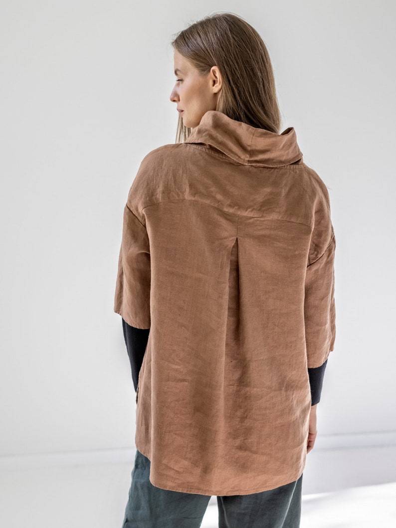May include: A brown linen shirt with a high collar and three-quarter sleeves. The shirt has a relaxed fit and a button-down front. The sleeves are dark blue. The model is wearing dark green pants.