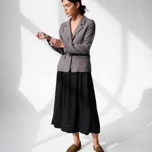 May include: A woman wearing a gray blazer over a black skirt and brown loafers. The blazer has a two-button closure and a notched lapel. The skirt is a midi length, and the loafers are suede.
