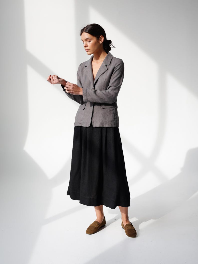 May include: A woman wearing a gray blazer over a black skirt and brown loafers. The blazer has a two-button closure and a notched lapel. The skirt is a midi length, and the loafers are suede.