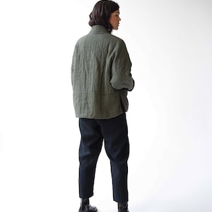 May include: An olive green quilted jacket with a high collar and black trousers. The jacket has a textured appearance and a relaxed fit. The trousers are a solid black colour. The person is wearing black boots.