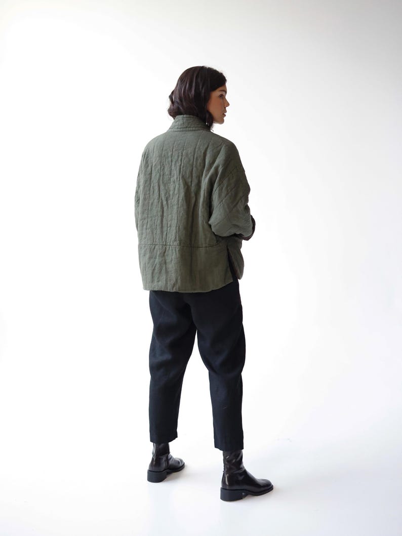 May include: An olive green quilted jacket with a high collar and black trousers. The jacket has a textured appearance and a relaxed fit. The trousers are a solid black colour. The person is wearing black boots.