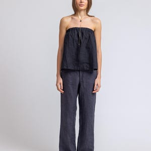 May include: A dark navy blue strapless top and matching wide-leg pants. The top has a gathered neckline and a gold-colored necklace. The pants are a solid color and have a relaxed fit. The outfit is made of a lightweight fabric.