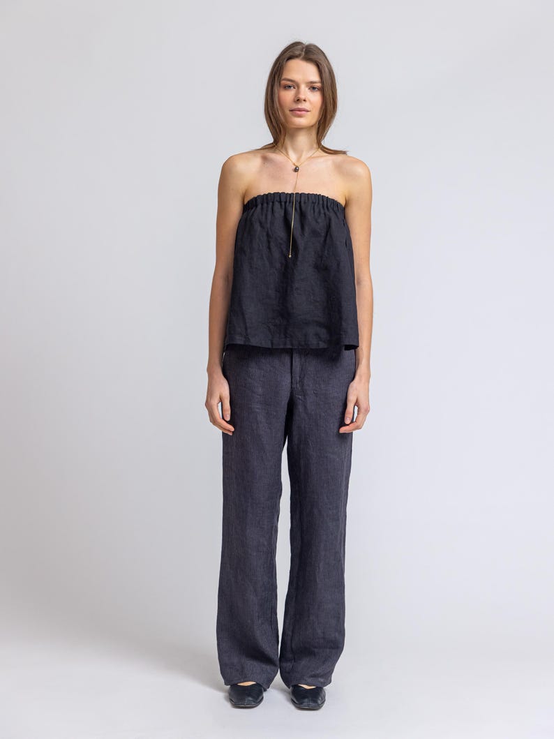 May include: A dark navy blue strapless top and matching wide-leg pants. The top has a gathered neckline and a gold-colored necklace. The pants are a solid color and have a relaxed fit. The outfit is made of a lightweight fabric.