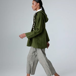 K&ouml;nnte beinhalten: Olivgr&uuml;ne Kapuzenjacke, wei&szlig;es Hemd und graue Leinenhose, kombiniert mit wei&szlig;en Turnschuhen. Das Outfit ist l&auml;ssig und bequem. Das Model geht. Die Hose ist hellgrau.
