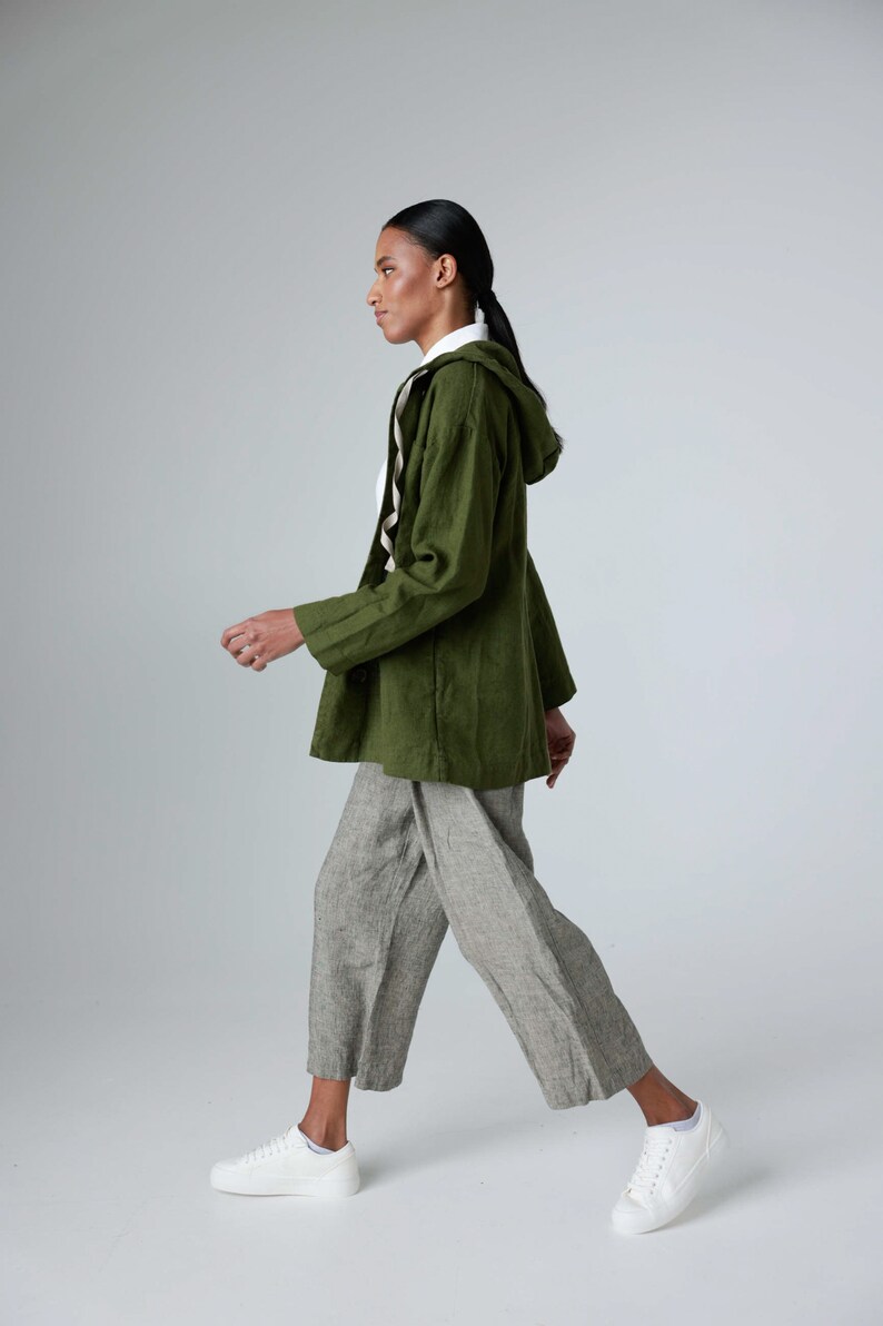 K&ouml;nnte beinhalten: Olivgr&uuml;ne Kapuzenjacke, wei&szlig;es Hemd und graue Leinenhose, kombiniert mit wei&szlig;en Turnschuhen. Das Outfit ist l&auml;ssig und bequem. Das Model geht. Die Hose ist hellgrau.