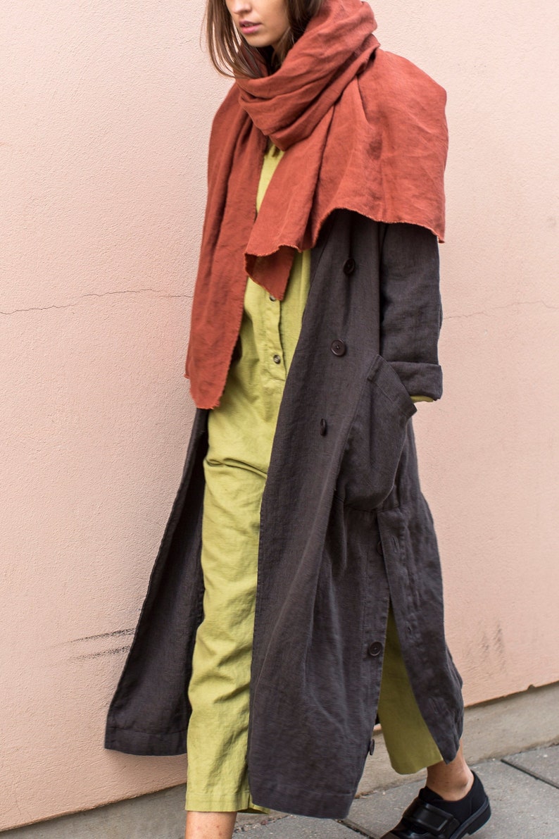 May include: A woman wearing a rust-colored scarf, a light green jumpsuit, and a dark gray long coat. The coat has large buttons and pockets. The outfit is a stylish ensemble.
