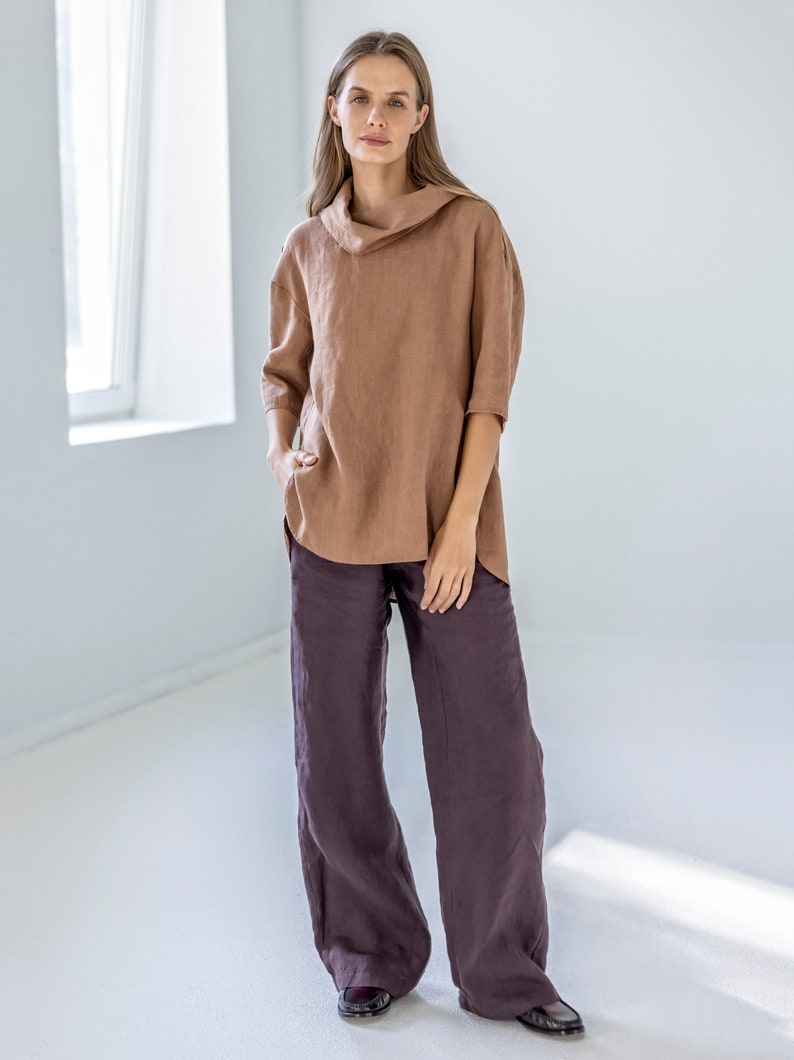 May include: A woman is wearing a brown linen top with a cowl neck and three-quarter sleeves, paired with wide-leg, dark purple linen pants. The outfit is simple and elegant, with a relaxed fit.