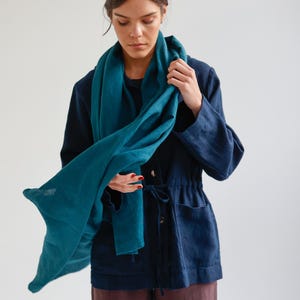 May include: A teal-colored scarf draped over a navy blue jacket. The scarf is made of a soft, flowing fabric. The jacket has a drawstring waist and two front pockets. The person is wearing dark brown pants.