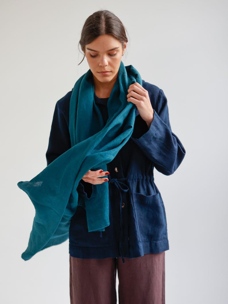 May include: A teal-colored scarf draped over a navy blue jacket. The scarf is made of a soft, flowing fabric. The jacket has a drawstring waist and two front pockets. The person is wearing dark brown pants.