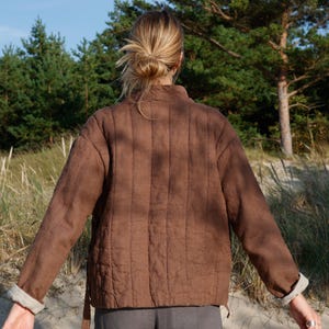 May include: A brown quilted jacket with a stand-up collar and long sleeves. The jacket has a textured appearance and a light-colored cuff detail. The person is wearing gray pants. The background includes trees and sand.