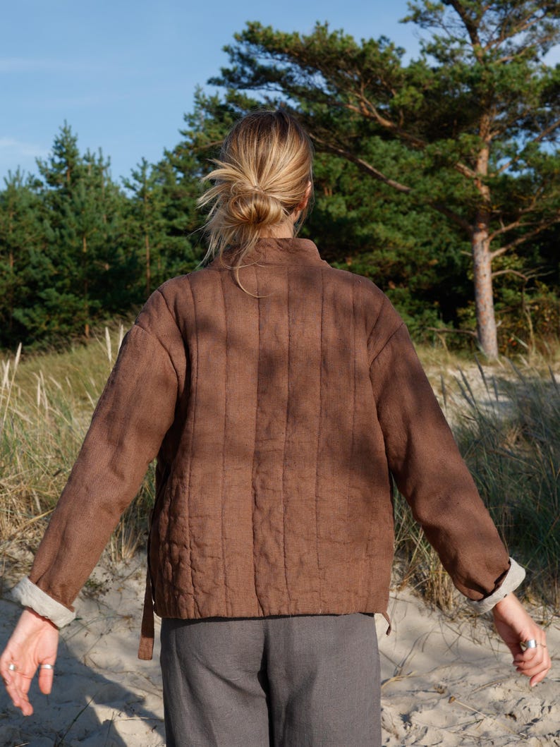May include: A brown quilted jacket with a stand-up collar and long sleeves. The jacket has a textured appearance and a light-colored cuff detail. The person is wearing gray pants. The background includes trees and sand.