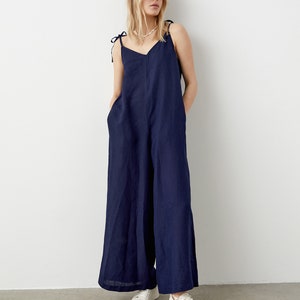 May include: A navy blue jumpsuit with wide legs and tie straps. The jumpsuit has a V-neck and pockets. The model is wearing white sneakers. The background is a plain white wall and a gray floor.