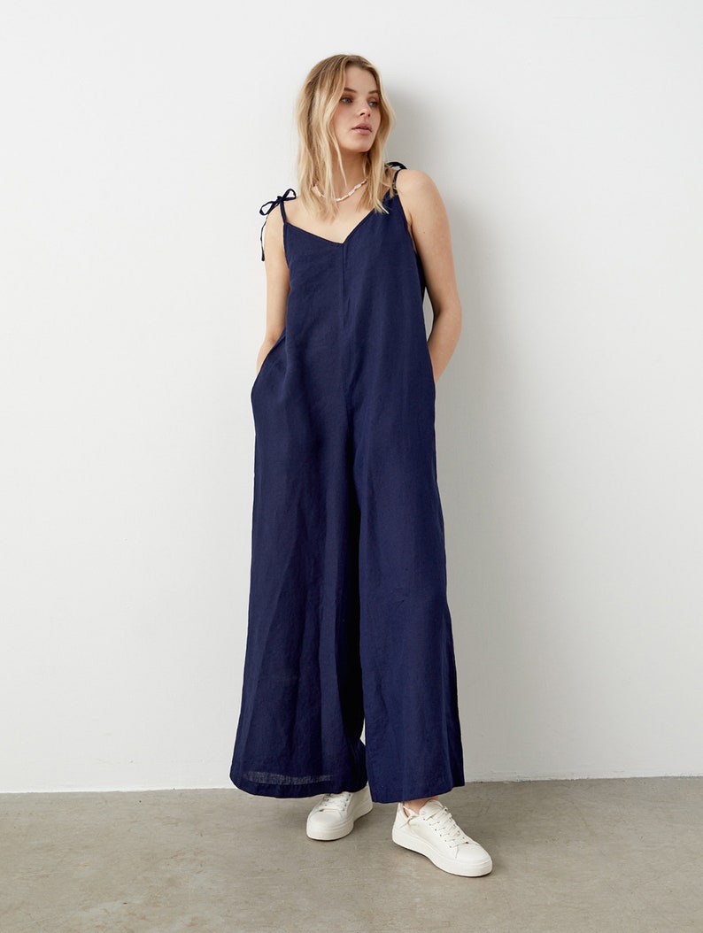 May include: A navy blue jumpsuit with wide legs and tie straps. The jumpsuit has a V-neck and pockets. The model is wearing white sneakers. The background is a plain white wall and a gray floor.