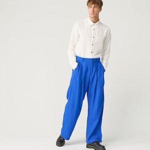 May include: A person wearing a white long-sleeved button-down shirt and blue pleated pants. They are standing with their hands in their pockets and looking to the side.