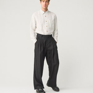 Op de afbeelding: Een persoon draagt een lichtgekleurd, verticaal gestreept overhemd met houten knopen en een donkere, wijd uitlopende broek. De outfit wordt gecompleteerd met zwarte leren schoenen. De kleding is casual en stijlvol.