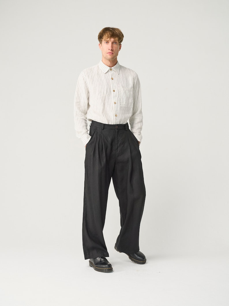 Op de afbeelding: Een persoon draagt een lichtgekleurd, verticaal gestreept overhemd met houten knopen en een donkere, wijd uitlopende broek. De outfit wordt gecompleteerd met zwarte leren schoenen. De kleding is casual en stijlvol.