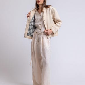 May include: A three-piece linen outfit in a neutral color. The outfit includes a jacket with a gingham lining, a button-down shirt, and wide-leg pants. The jacket has dark buttons, and the pants have a matching belt. The model is wearing white sandals.