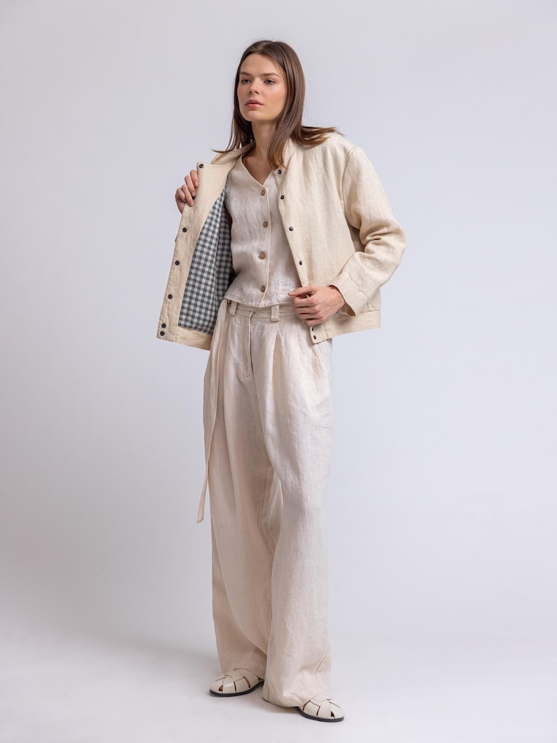 May include: A three-piece linen outfit in a neutral color. The outfit includes a jacket with a gingham lining, a button-down shirt, and wide-leg pants. The jacket has dark buttons, and the pants have a matching belt. The model is wearing white sandals.