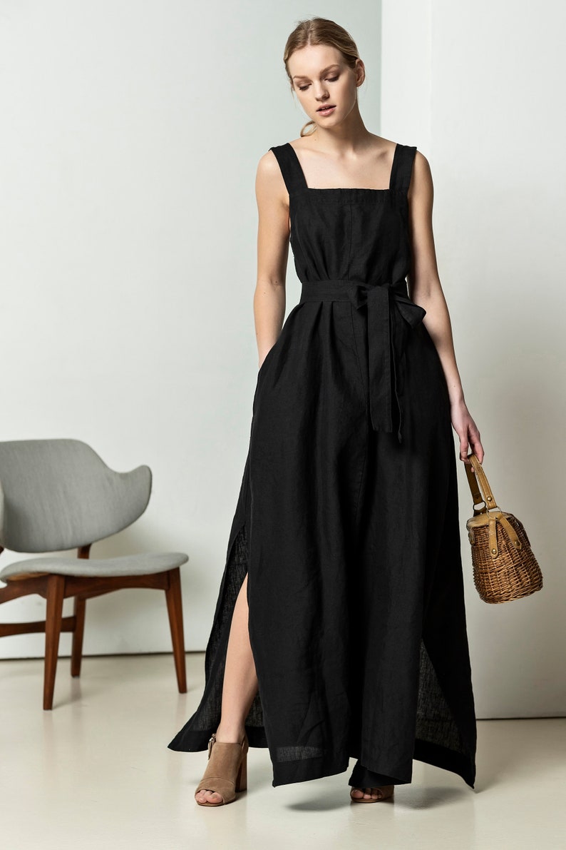 May include: A black linen maxi dress with a square neckline, sleeveless design, and a self-tie belt. The dress has side slits and is shown with a brown wicker handbag.