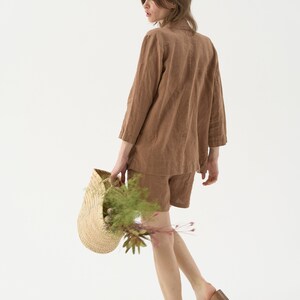 May include: A woman wearing a brown linen blazer and shorts set. She is carrying a woven straw basket filled with greenery and flowers. The outfit is a neutral color, and she is wearing sandals.
