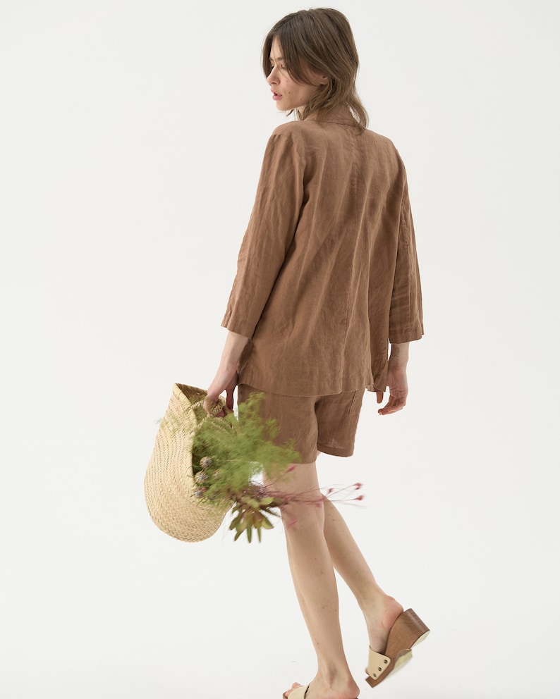 May include: A woman wearing a brown linen blazer and shorts set. She is carrying a woven straw basket filled with greenery and flowers. The outfit is a neutral color, and she is wearing sandals.