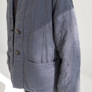 May include: A slate blue quilted jacket with a relaxed fit. The jacket has a V-neck, two large wooden buttons, and two front pockets. The fabric has vertical stitching details and a slightly worn appearance, suggesting a vintage or handcrafted style.