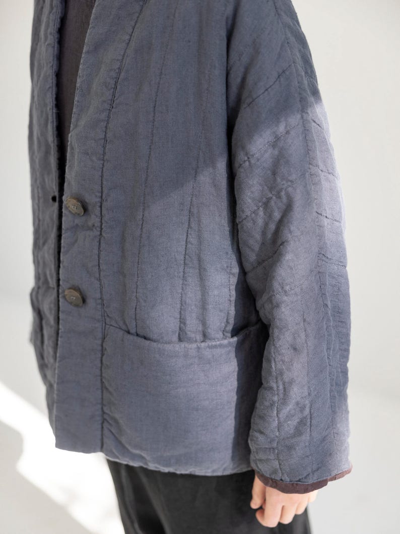 May include: A slate blue quilted jacket with a relaxed fit. The jacket has a V-neck, two large wooden buttons, and two front pockets. The fabric has vertical stitching details and a slightly worn appearance, suggesting a vintage or handcrafted style.