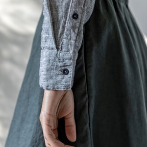 May include: A close-up of a gray and white checkered long-sleeve shirt with dark blue buttons and a dark green skirt. The fabric appears to be linen or a similar lightweight material. The image shows a person's arm and hand.