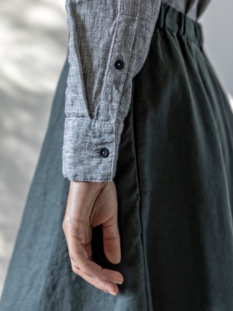 May include: A close-up of a gray and white checkered long-sleeve shirt with dark blue buttons and a dark green skirt. The fabric appears to be linen or a similar lightweight material. The image shows a person's arm and hand.