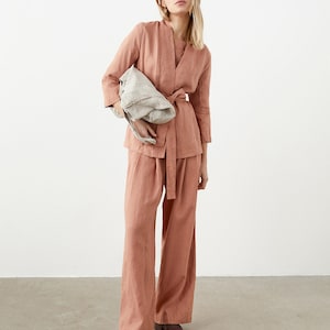 May include: A salmon-colored linen outfit consisting of a wrap top with a matching belt and wide-leg pants. The outfit is paired with a light gray tote bag and brown shoes. The ensemble is made of a lightweight fabric, perfect for a relaxed, yet stylish look.
