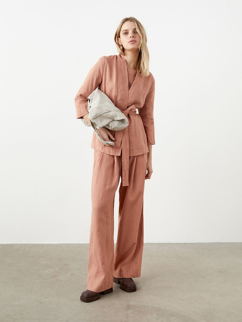 May include: A salmon-colored linen outfit consisting of a wrap top with a matching belt and wide-leg pants. The outfit is paired with a light gray tote bag and brown shoes. The ensemble is made of a lightweight fabric, perfect for a relaxed, yet stylish look.
