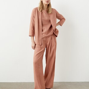 May include: A three-piece linen outfit in a muted coral color. The ensemble includes a long-sleeved top, a matching open-front jacket, and wide-leg pants. The outfit is made of a lightweight fabric, suitable for warm weather.
