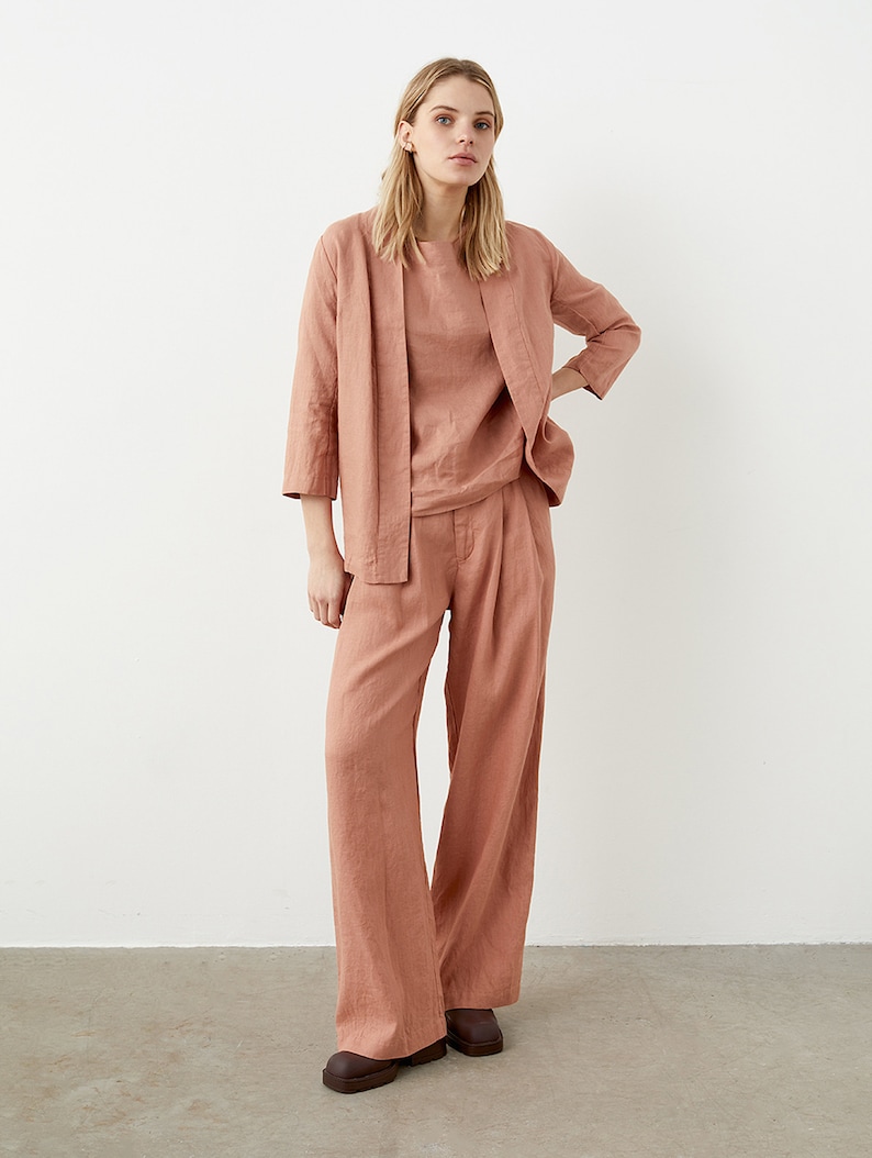 May include: A three-piece linen outfit in a muted coral color. The ensemble includes a long-sleeved top, a matching open-front jacket, and wide-leg pants. The outfit is made of a lightweight fabric, suitable for warm weather.