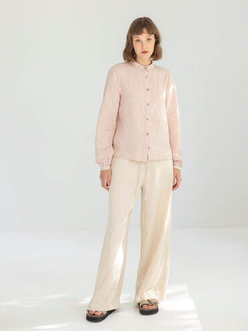 May include: A light pink linen shirt with a mandarin collar and wooden buttons is paired with cream-colored linen pants. The pants have a relaxed fit and a drawstring waist. The outfit is completed with black sandals.
