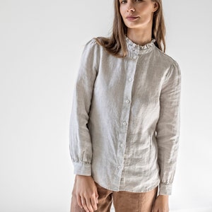 Può includere: Camicia grigio chiaro con un piccolo motivo a quadri e colletto a balze. La camicia a maniche lunghe ha un design classico con bottoni sul davanti ed è abbinata a pantaloni marroni.