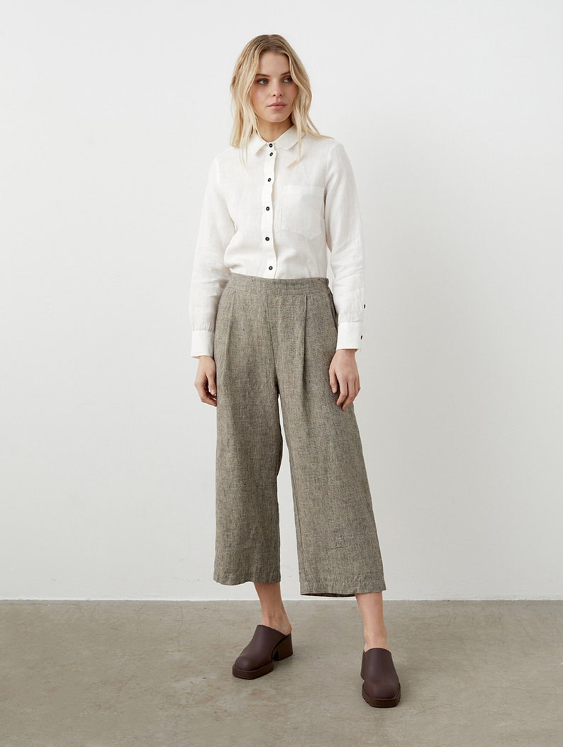 K&ouml;nnte beinhalten: Eine Frau tr&auml;gt ein wei&szlig;es Hemd mit schwarzen Kn&ouml;pfen und eine olivgr&uuml;ne, weit geschnittene, cropped Hose. Sie tr&auml;gt auch braune Lederclogs. Das Outfit ist schlicht und elegant.