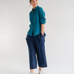 May include: A teal button-down shirt with rolled-up sleeves and navy blue culottes. The outfit is paired with white sneakers. The clothing appears to be made of linen or a similar lightweight fabric, suitable for warm weather.