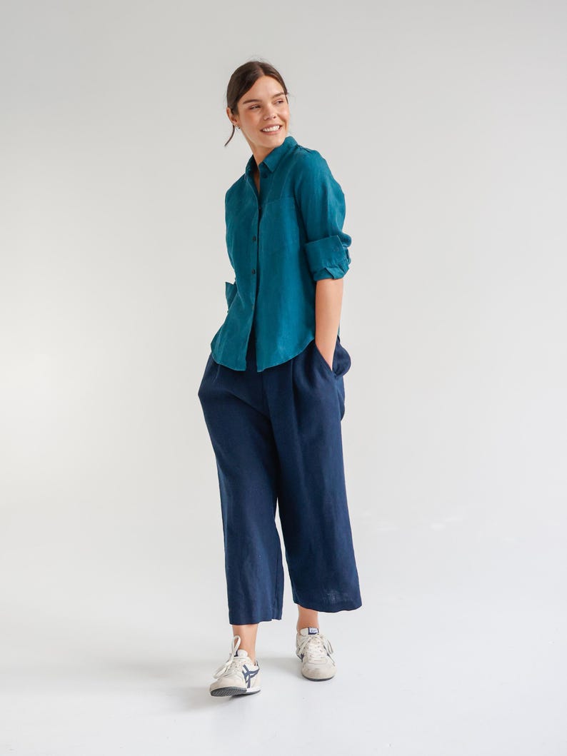 May include: A teal button-down shirt with rolled-up sleeves and navy blue culottes. The outfit is paired with white sneakers. The clothing appears to be made of linen or a similar lightweight fabric, suitable for warm weather.