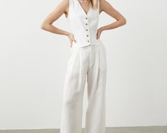 Pode incluir: Um conjunto branco de linho composto por um colete com botões de madeira e calças largas a condizer. O conjunto é complementado com sapatilhas pretas. O conjunto é de cor neutra e parece ser feito de um tecido leve, adequado para o clima quente.