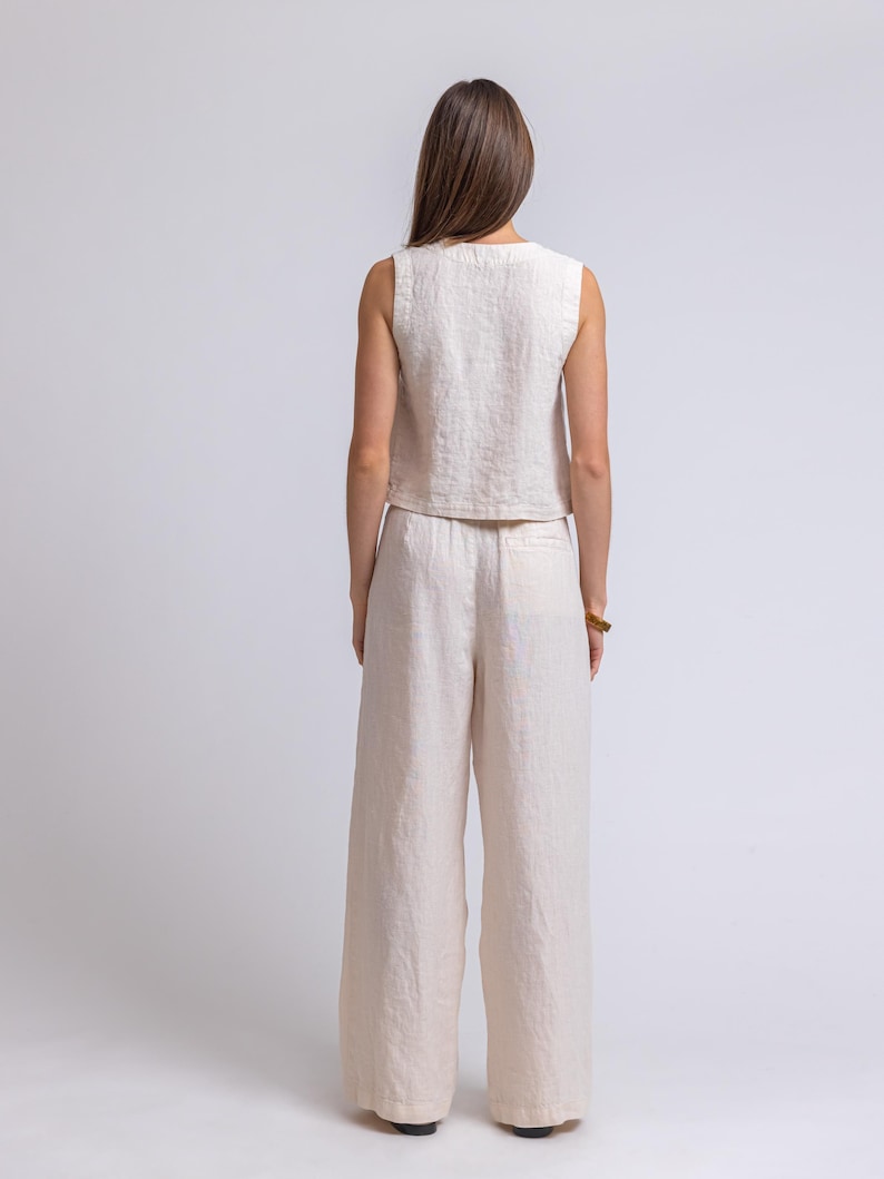 May include: A sleeveless, cream-colored linen top and wide-leg pants set. The outfit is made of a textured fabric and has a relaxed fit. The model is facing away from the camera, showcasing the back of the outfit.