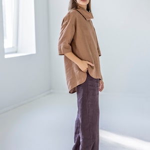 May include: A woman wearing a brown linen top with a wide collar and three-quarter sleeves, paired with dark purple wide-leg linen pants. The outfit is simple and elegant, with a focus on natural fabrics and a relaxed fit.