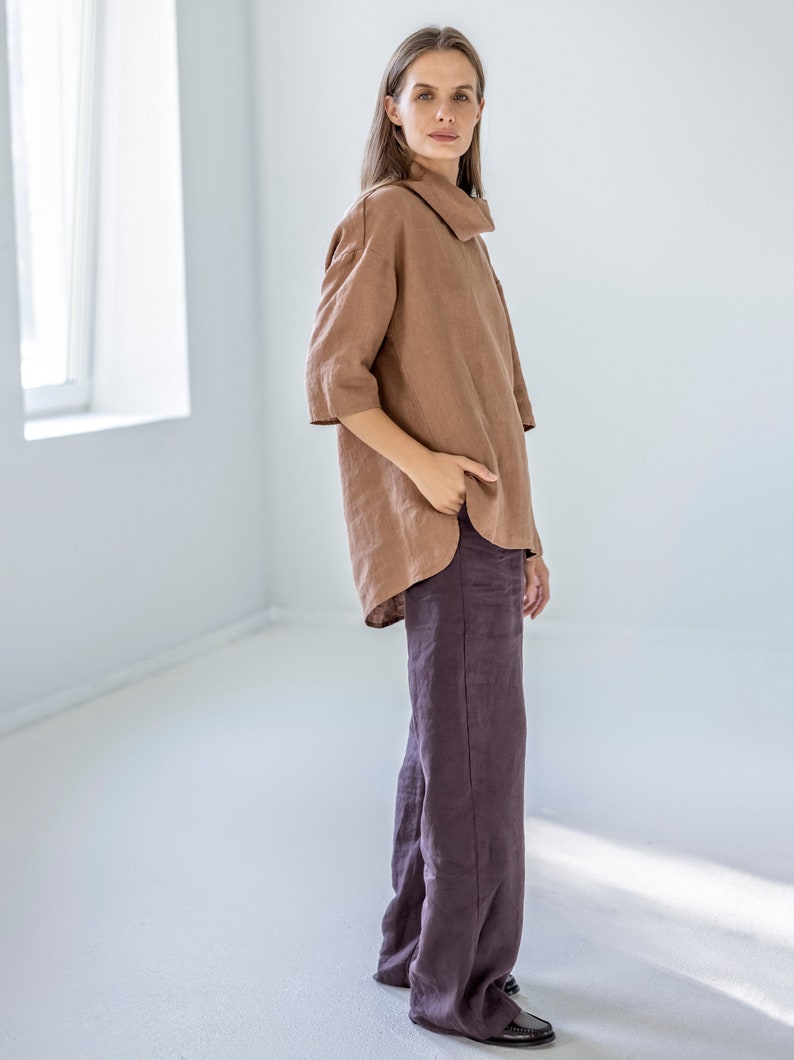 May include: A woman wearing a brown linen top with a wide collar and three-quarter sleeves, paired with dark purple wide-leg linen pants. The outfit is simple and elegant, with a focus on natural fabrics and a relaxed fit.