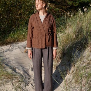 May include: A woman wearing a brown quilted jacket with a tie closure and gray linen pants. The jacket has a diamond pattern and long sleeves. The pants are wide-legged. The outfit is worn on a sandy beach with tall grass and trees in the background.