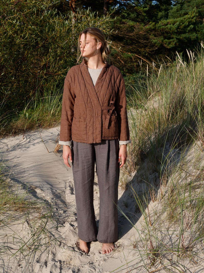 May include: A woman wearing a brown quilted jacket with a tie closure and gray linen pants. The jacket has a diamond pattern and long sleeves. The pants are wide-legged. The outfit is worn on a sandy beach with tall grass and trees in the background.