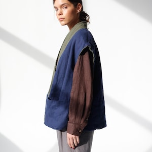 May include: A layered outfit featuring a blue vest with a green collar, a brown long-sleeve shirt, and gray pants. The vest has large pockets. The model is facing the camera, with natural light illuminating the scene.