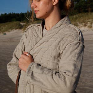 May include: A light beige quilted jacket with a wrap-style closure. The jacket has a brown trim and is worn by a person on a beach. The jacket's fabric appears to be linen or a similar natural material. The sleeves are long.