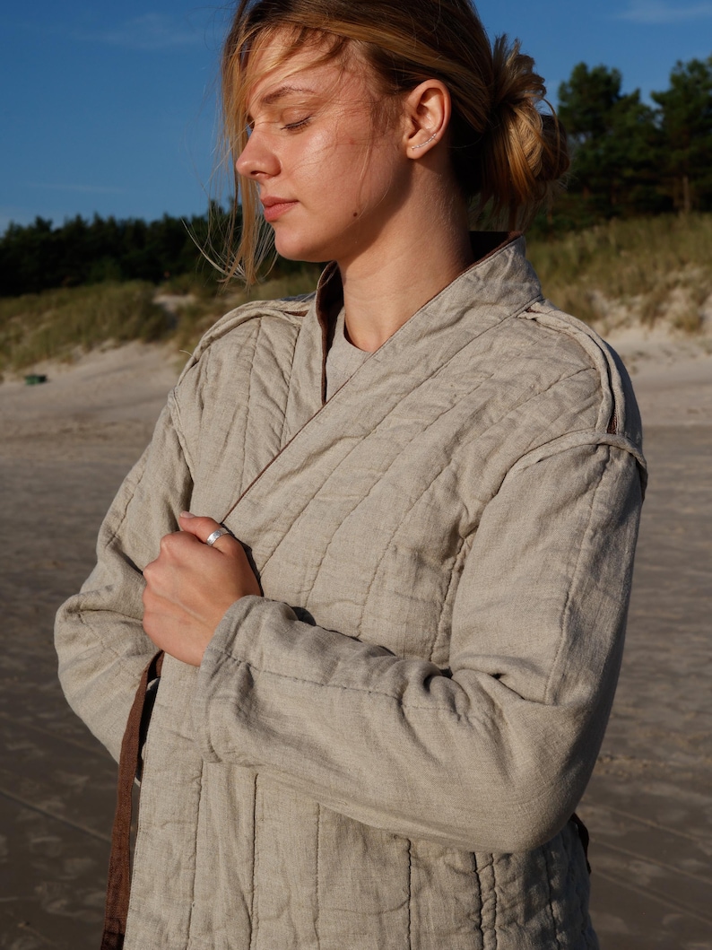 May include: A light beige quilted jacket with a wrap-style closure. The jacket has a brown trim and is worn by a person on a beach. The jacket's fabric appears to be linen or a similar natural material. The sleeves are long.