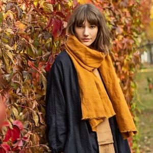 May include: A woman wearing a mustard yellow scarf with a fringed edge, a navy blue coat, and a tan top. The background features colorful autumn foliage with red, orange, and green leaves.