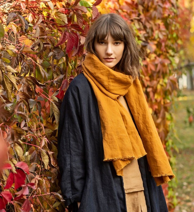 May include: A woman wearing a mustard yellow scarf with a fringed edge, a navy blue coat, and a tan top. The background features colorful autumn foliage with red, orange, and green leaves.